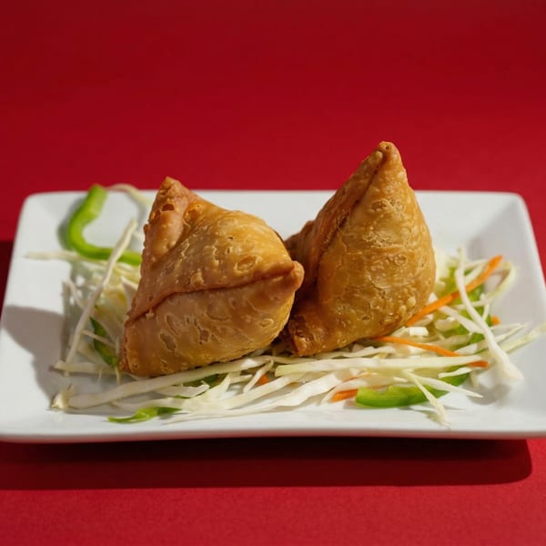 Two golden-brown samosas rest on a bed of shredded cabbage and bell peppers on a white plate.