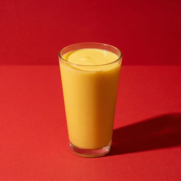 A tall glass of bright yellow mango lassi sits on a vibrant red surface.